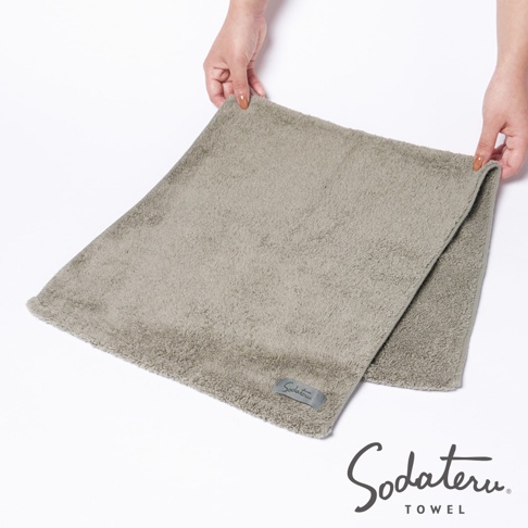 一條淺灰色、蓬鬆柔軟的毛巾，材質為厚實的棉絨，帶有品牌標籤。