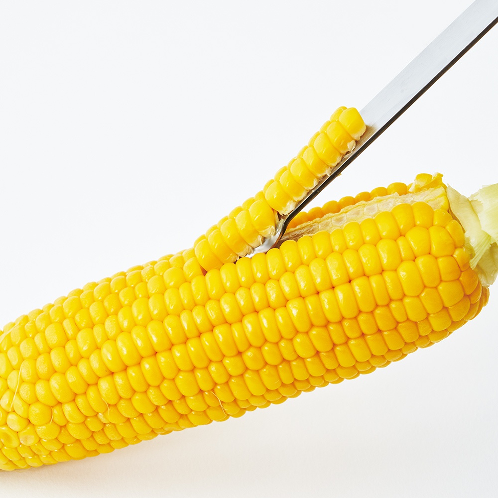 日製玉米刨刀正在將一根飽滿的黃色玉米的玉米粒刨下，玉米粒呈金黃色，排列整齊。