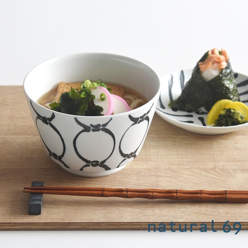 白色陶瓷丼碗，飾有黑色手繪迴紋圖案，內盛烏龍麵，配料有豆腐皮、海帶芽、魚板及蔥花。旁邊盤子裡有一個飯糰，旁邊有醃漬物和檸檬。