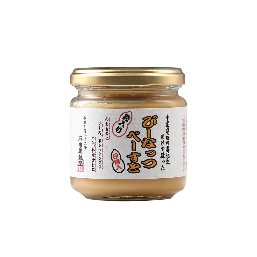 玻璃罐裝的顆粒花生醬，淺褐色醬體，頂部有金色金屬旋蓋，標籤為白色，印有日文說明，文字顏色包含黑色、紅色和橘色。