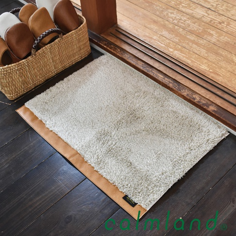 淺灰色絨面玄關地墊，邊緣有棕色皮革飾邊，放置於深色木地板上，旁有藤編籃子裝著室內拖鞋。