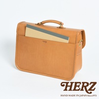 淺棕色皮革製成的方形單肩包，配有頂部提把和可拆卸肩帶。包身正面有一個大翻蓋口袋，內含一本筆記本。包包採用對比色縫線，並飾有「HERZ」壓花標誌。
