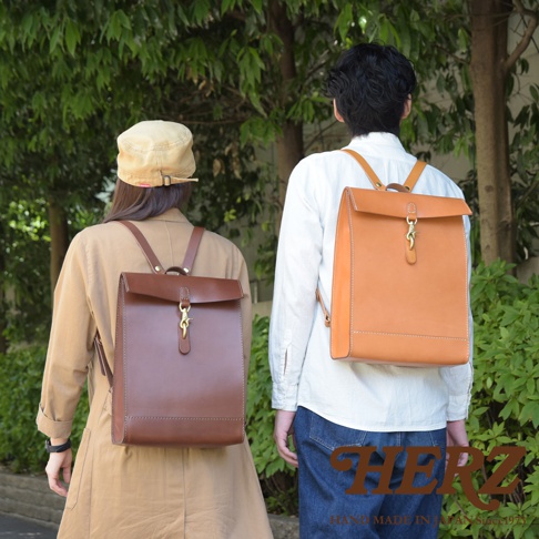 兩位模特兒背著職人手作皮革紙袋風後背包，一位背著深棕色，另一位背著淺棕色。背包設計簡約，材質為皮革，具有紙袋般的方正外型，並以金屬扣環開合。