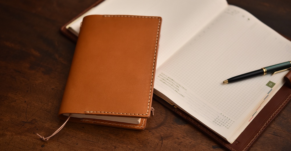 一個棕褐色的皮革手帳書套，旁邊攤開一本筆記本，筆記本上有網格線和日曆，一支綠色和金色的筆放在筆記本上，背景是木紋桌面。