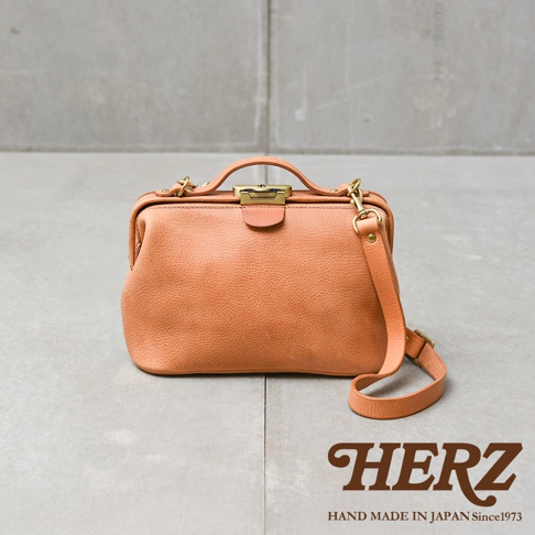 HERZ 品牌橘色復古皮革手提斜背包，配有金色鎖扣和可調式背帶，包款底部平整，呈現職人手作的精緻工藝。