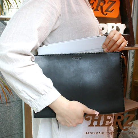 一位女士手持一個黑色皮革手拿包，包身簡約，頂部有拉鍊和品牌標誌。包內裝有一個淺灰色平板電腦。