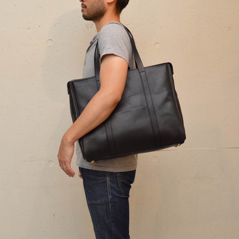 一名男子肩背一個黑色荔枝紋皮革托特包，包包有拉鍊開合，外部有縫線裝飾的插袋，底部有金屬腳釘。