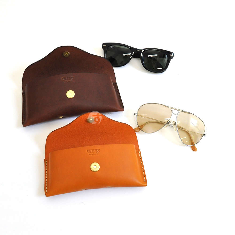 兩個皮革眼鏡收納盒，一個深棕色，一個橘棕色，旁邊擺著一副黑色太陽眼鏡和一副飛行員款式的太陽眼鏡。