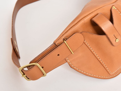 職人手作皮革斜背包，採用天然棕色皮革，帶有黃銅色金屬扣環和铆釘，手工縫線清晰，展現細膩質感。