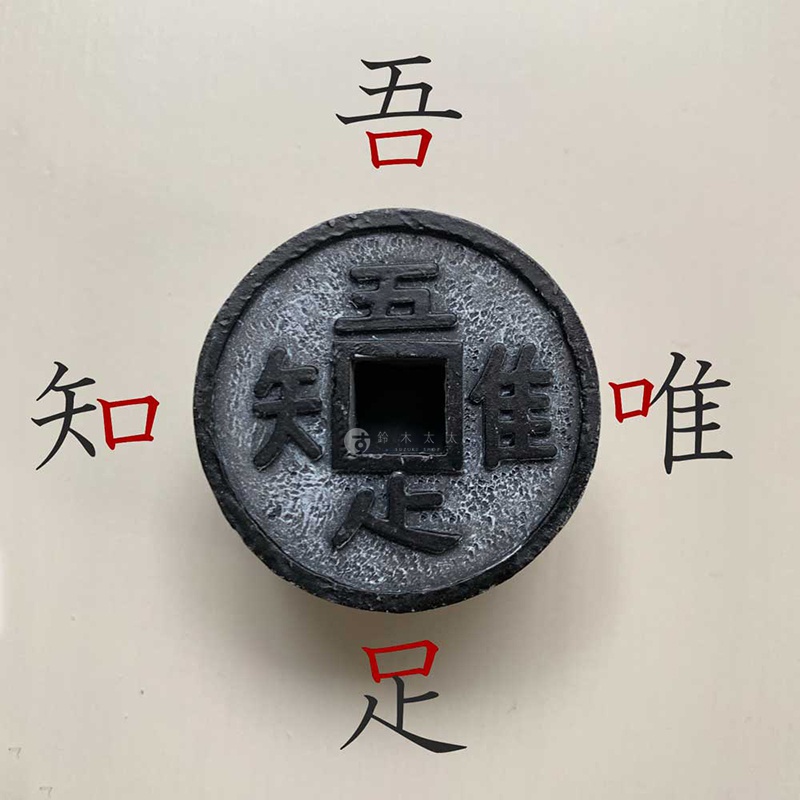 這是一個仿古圓形銅錢造型的擺件，中央設有方形孔洞，錢幣四邊刻有「吾、唯、知、足」四個漢字，表面呈現灰黑色帶有斑駁的復古金屬質感，整體置於淺色背景中央。