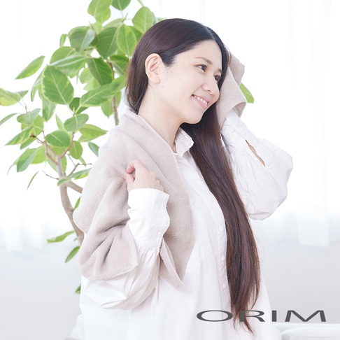 一位女子在綠植旁，頭上搭著一條米色浴巾，正在用另一條浴巾擦拭臉頰，浴巾質地柔軟，呈現出細緻的紋理。