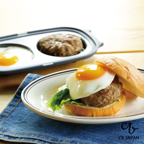 CB JAPAN copan系列 微波爐專用燒烤盤，在瓷盤上放著一個漢堡，漢堡上方蓋著一顆煎好的太陽蛋，盤子旁有一塊藍色牛仔布，背景是木紋桌面和一個微波爐專用燒烤盤，燒烤盤裡有兩個煎好的荷包蛋和一個漢堡排。