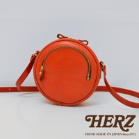 橘色皮革小圓包，圓形包身，正面有半圓形拉鍊開口，搭配金色拉鍊及同色系皮革拉頭，附可調式皮革背帶。