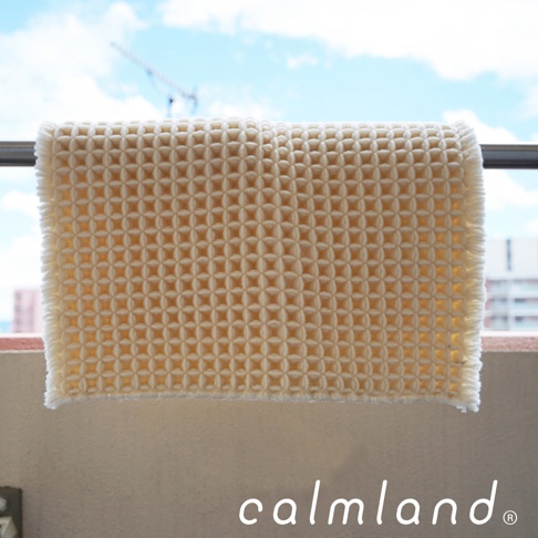 淺黃色鬆餅紋瞬乾吸水浴墊，材質柔軟，邊緣有流蘇，懸掛在戶外，背景為藍天白雲與建築物。