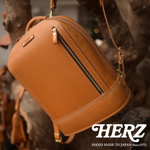 一個由棕色皮革製成的立體商務後背包，具有明顯的縫線和金屬拉鍊，背包懸掛在樹枝上，背景模糊。