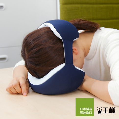 深藍色與白色相間的「王樣の舒眼枕」，以柔軟材質包裹住一位女性的頭部，提供頸部支撐，臉部被枕頭輕柔覆蓋，雙手搭在桌面上，呈現舒適的午休姿態。