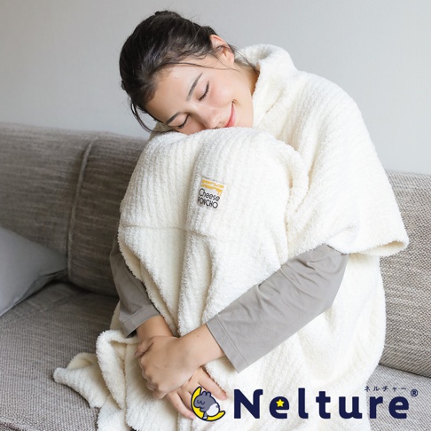 一位年輕女子蜷縮在沙發上，身上裹著一件奶油色的極柔毛絨起司斗篷，臉上帶著滿足的微笑。斗篷上印有「Cheese Poncho」字樣，下方還有「Nelture」品牌標誌。