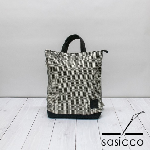 三河木綿刺子織手提後背包，以黑白格紋圖案為主體，搭配黑色底部與提把，側面有一個黑色方形布標。