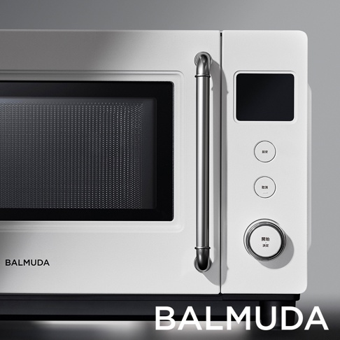 BALMUDA 品牌白色微波爐的近拍特寫，畫面呈現產品右側的控制面板、金屬質感把手與黑色烤漆門板，面板上有三個圓形按鈕與一個顯示螢幕，下方文字標示 BALMUDA。