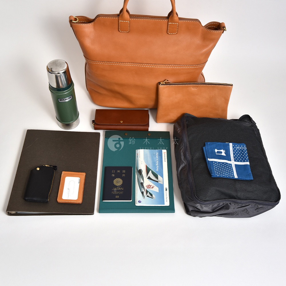 一只淺棕色皮革托特包，搭配一個同色系拉鍊小包、一個棕色皮夾、一個綠色保溫瓶、一個黑色證件夾、一個卡夾、一本綠色文件夾、一本護照、一支筆以及一個黑色收納袋，袋內有一塊藍色印花手帕。