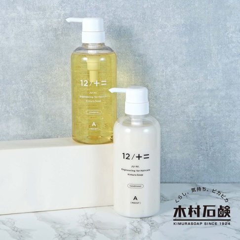 兩瓶 JU-NI 潤髮乳，一瓶為透明金黃色液體，另一瓶為乳白色乳液，皆為白色壓頭的透明塑膠瓶裝，瓶身印有黑色 LOGO 和產品資訊，背景為灰色紋理牆面。