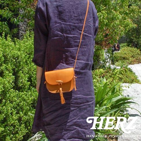 職人手作皮革流蘇側背包，橘棕色皮革，帶有木質鈕扣和皮革流蘇裝飾，斜挎於身穿深紫色長裙的模特兒身上，背景為綠色植物與戶外景觀。