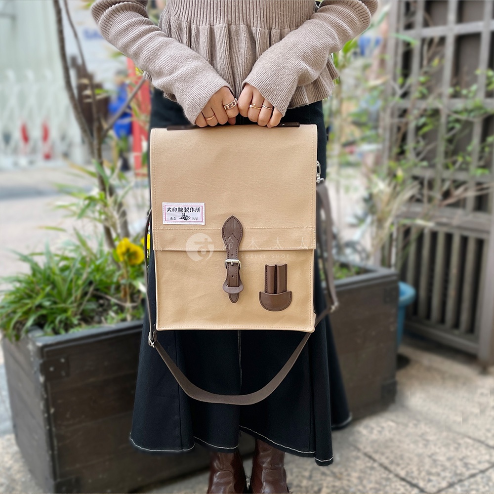 米色純棉帆布製成，綴有棕色皮革飾邊的直式側背包。包款有皮帶扣和兩個皮革插槽設計，配有可拆卸的肩帶。