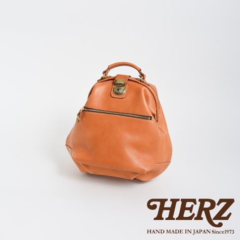 職人手作皮革輕旅行後背包，採用溫潤的淺棕色皮革，帶有復古風格的銅色金屬鎖扣和拉鍊，包身線條圓潤，質感厚實，適合日常或短途旅行攜帶。