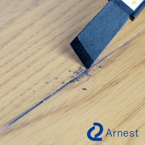 黑色清潔刮刀的尖端正在清理木地板縫隙中的灰色污垢，刮刀下方留有少量細碎的黑色顆粒。