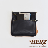 黑色皮革翻轉直式斜背包，正面有一個拉鍊口袋，內含手機與書籍，背包邊緣露出天然皮革材質。