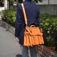 職人手作皮革旅行包 A-102，採用橘棕色皮革，設計有兩個前袋、頂部提把和可調式肩帶，搭配金屬扣環與縫線細節，適合肩背或斜背。