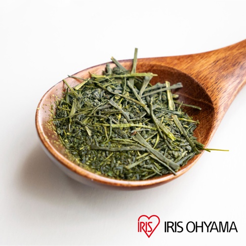 木製湯匙盛裝著乾燥的綠茶葉，深綠色針狀茶葉與細碎茶末清晰可見，湯匙右下角標有紅色心形圖案的IRIS OHYAMA品牌標誌，背景為簡潔的白色。