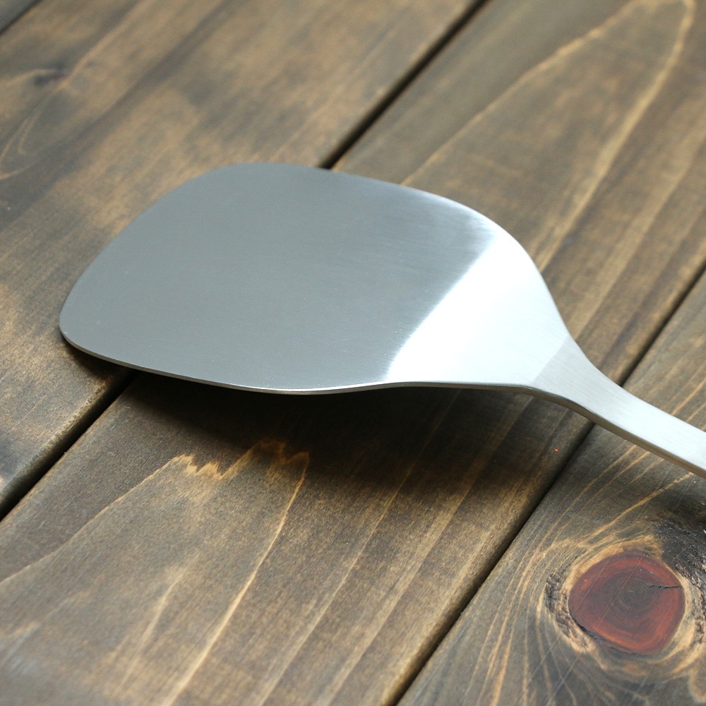 銀色不鏽鋼萬用鍋鏟，寬扁的鏟面，有著細緻的拉絲紋理，放在深棕色木紋桌面上。