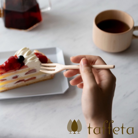 一隻手拿著一支淺色木製點心叉，叉子有三齒，靠近一塊裝飾有鮮奶油、草莓和藍莓的蛋糕。背景是淺色桌面，一旁放著一個木質咖啡杯和一個玻璃茶壺。