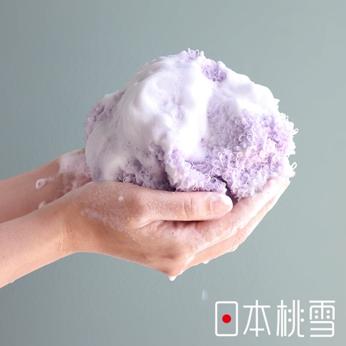 一雙手捧著一個淡紫色、充滿綿密泡沫的沐浴巾，沐浴巾呈現蓬鬆的球狀，材質為細緻的織紋，手部沾有泡沫。