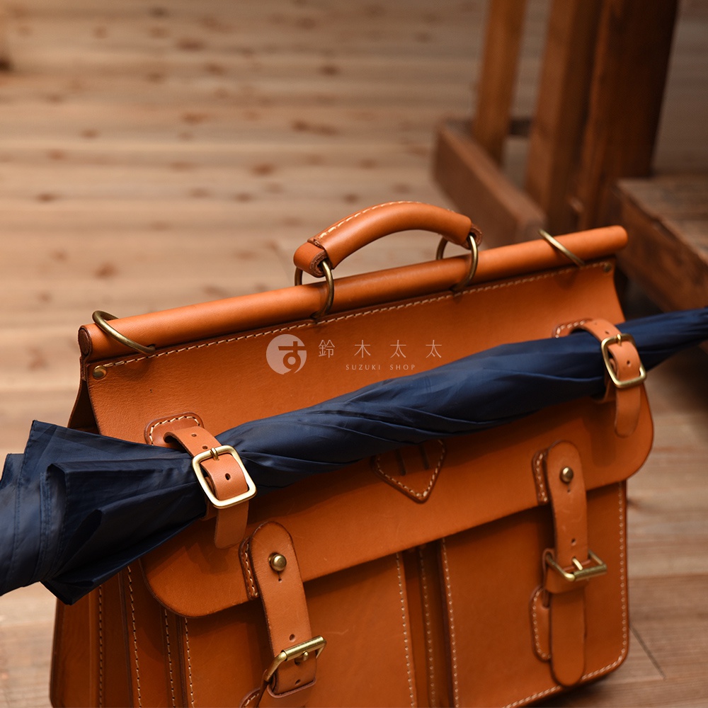 職人手作皮革旅行包，經典棕色，採用牛皮材質，配有金屬釦環與皮帶固定深藍色摺疊傘。