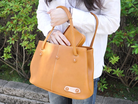 一位女士手提一個焦糖色皮革托特包，包包上有白色縫線裝飾，中間有兩顆圓形金屬扣。包包旁有一同色系的長夾。
