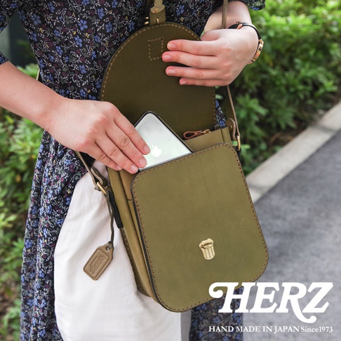一位穿著花卉洋裝的女性正將手機放入日本HERZ品牌的橄欖綠色硬皮革翻蓋迷你單肩包中，包包帶有復古金屬鎖扣與吊牌，呈現職人手作的高質感風格。
