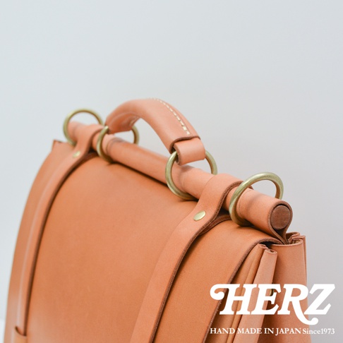 HERZ 品牌標誌與「HAND MADE IN JAPAN Since1973」字樣，輔以手工皮革側背包的細節特寫，呈現質感。