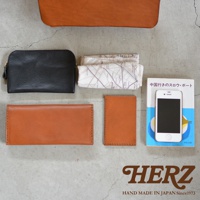 黑色皮革化妝包、茶色長夾、茶色名片夾、印有地圖圖案的布料、一本藍色封面的日文書籍和一部白色手機，擺放在灰色紋理的表面上，下方還有HERZ品牌標誌。