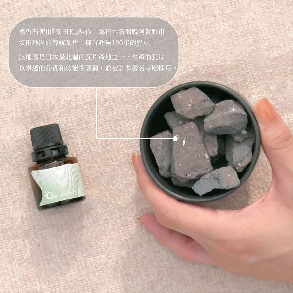 觸感粗糙的炭灰色擴香石散落在一個黑色圓碗中，由一隻手捧著，旁邊擺放著一小瓶深棕色精油，瓶身上有綠色漸層標籤。