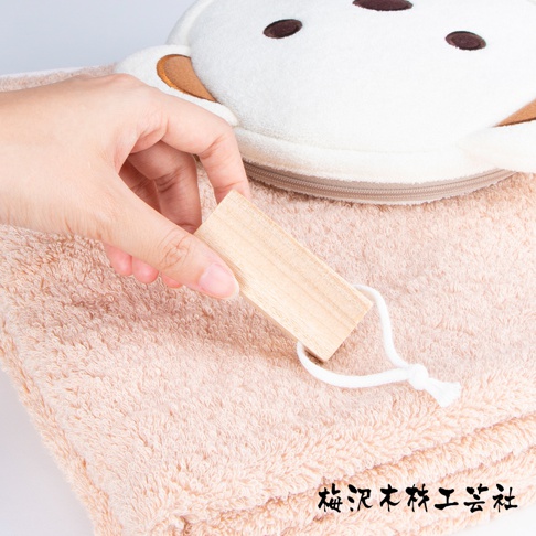 一隻手拿著一塊淺木色的方形樟木條，木條以白色細繩綁著，置於一條淺粉色、柔軟的毛巾上。毛巾上方有一個白色、帶有可愛熊臉圖案的絨布包。