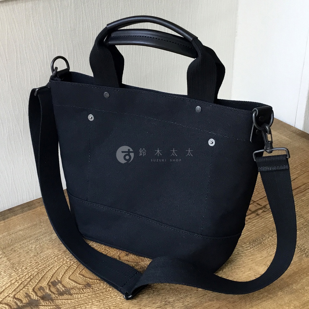 黑色帆布製成的兩用托特包，配有黑色皮革手柄和可拆卸斜挎肩帶，包身有銀色鉚釘裝飾，中央印有品牌標誌。