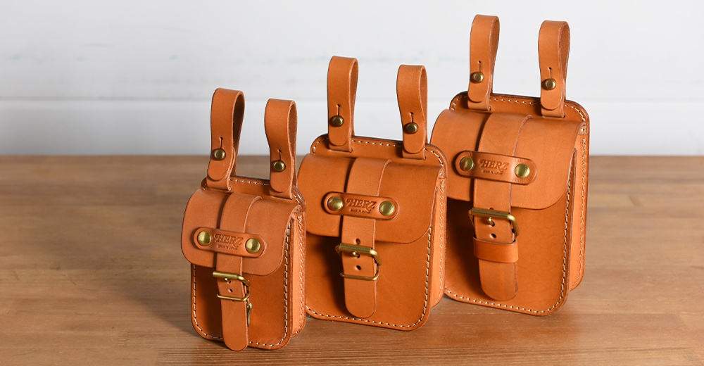 三款不同尺寸的職人手作皮革腰袋，皆為經典棕色，以手工縫製，設有皮帶扣和金屬釦環，並附有皮製懸掛帶。