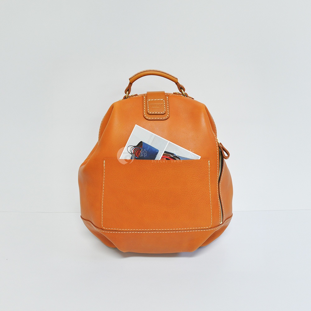 職人手作皮革輕旅行後背包，採用柔軟的橘棕色皮革製成，設計簡約。包包前方有一個外露口袋，內有雜誌或卡片等物品。包包頂部有提把，側邊有拉鍊。