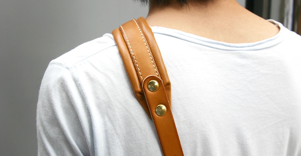職人手作皮革加厚減壓肩背帶，採用淺棕色皮革，搭配金色鉚釘，縫線清晰，展示於白色T恤上。