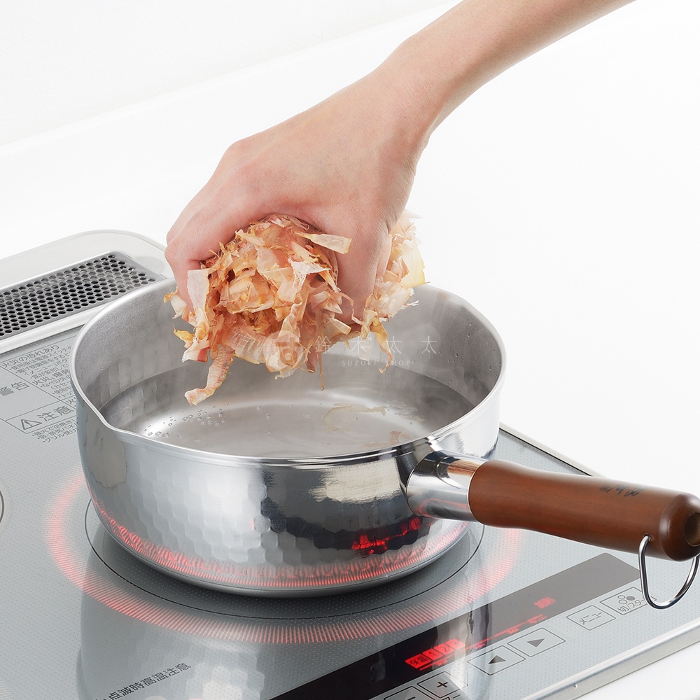 一個鄉技不鏽鋼單柄雪平鍋，鍋身有錘紋，搭配木質手柄，鍋內裝有水，一隻手正將柴魚片放入鍋中。