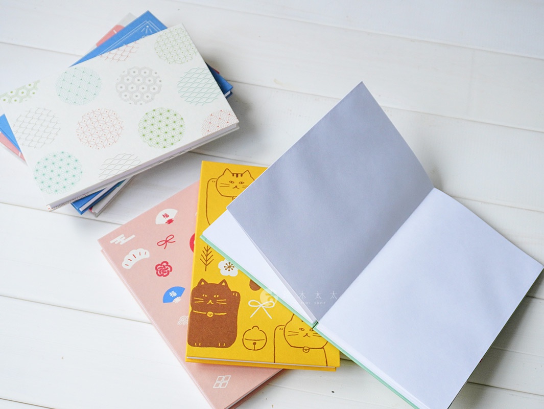 一疊美濃和紙御朱印帳，封面分別印有日式傳統圖案、粉色和風圖案及可愛的招財貓圖案。其中一本黃色封面御朱印帳攤開，露出內頁。