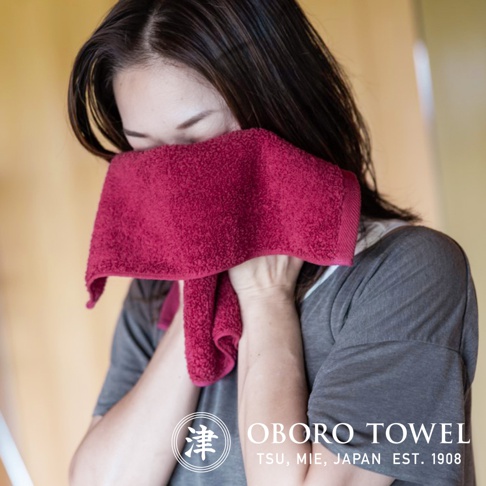 圖片中的文字為「津 ORO TOWEL TSU, MIE, JAPAN EST. 1908」。一位女子用一條紅色的毛巾擦拭臉部。