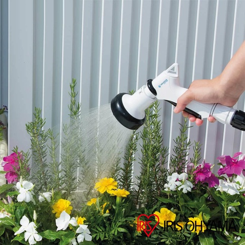 手持白色蓮蓬頭，對著一叢綠葉和五顏六色的花朵噴灑細水流，蓮蓬頭上有藍色標誌。